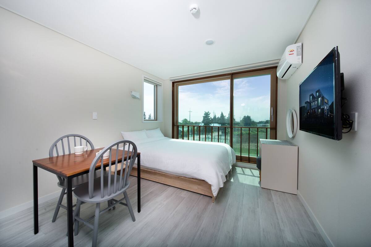 A modern hotel room with a bed, a dining table with chairs, a large window with a view of greenery, and a mounted television.