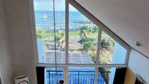 Une pièce dotée d’une grande fenêtre donnant sur la côte, d’un ventilateur de plafond, d’une petite table et d’un canapé.
