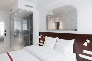 Una camera d'albergo moderna con un letto grande, un armadio a tutta parete in vetro, una toeletta con specchio e un grande specchio ad arco sopra il letto.