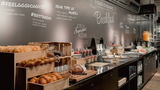 A breakfast buffet with various bread and pastries, a coffee station, and a wall with organic and vegan options. - Ruby Coco Hotel Dusseldorf by IHG (Düsseldorf)