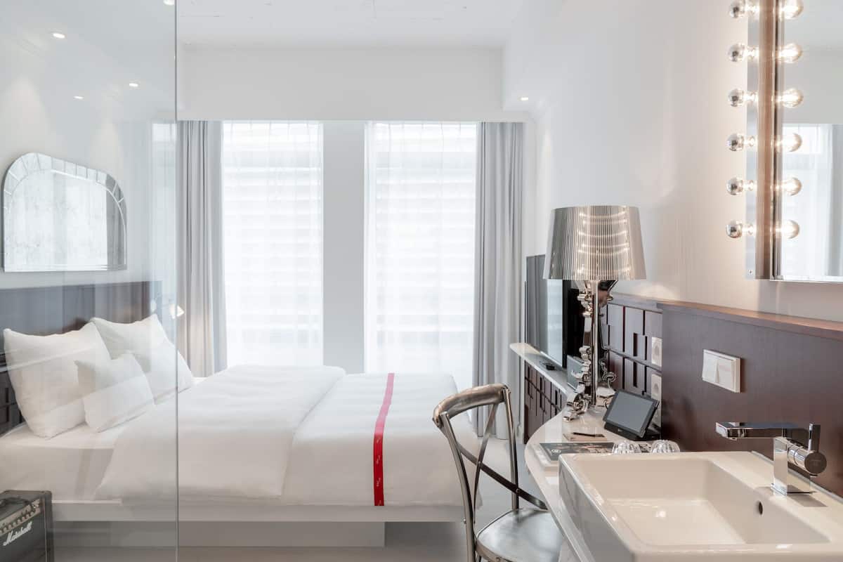 Habitación de hotel con cama, escritorio con computadora, espejo grande con luces y baño con lavamanos y espejo.