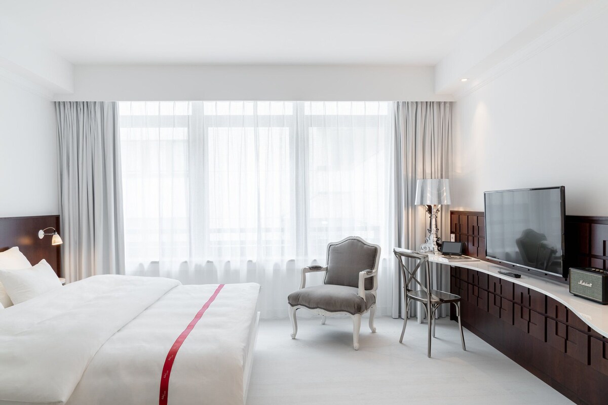 A hotel room with a bed, a desk with a chair, a television, and a large window with curtains.