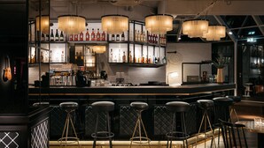 A bar area with multiple stools, a counter with bottles, and pendant lighting. - Ruby Coco Hotel Dusseldorf by IHG (Düsseldorf)