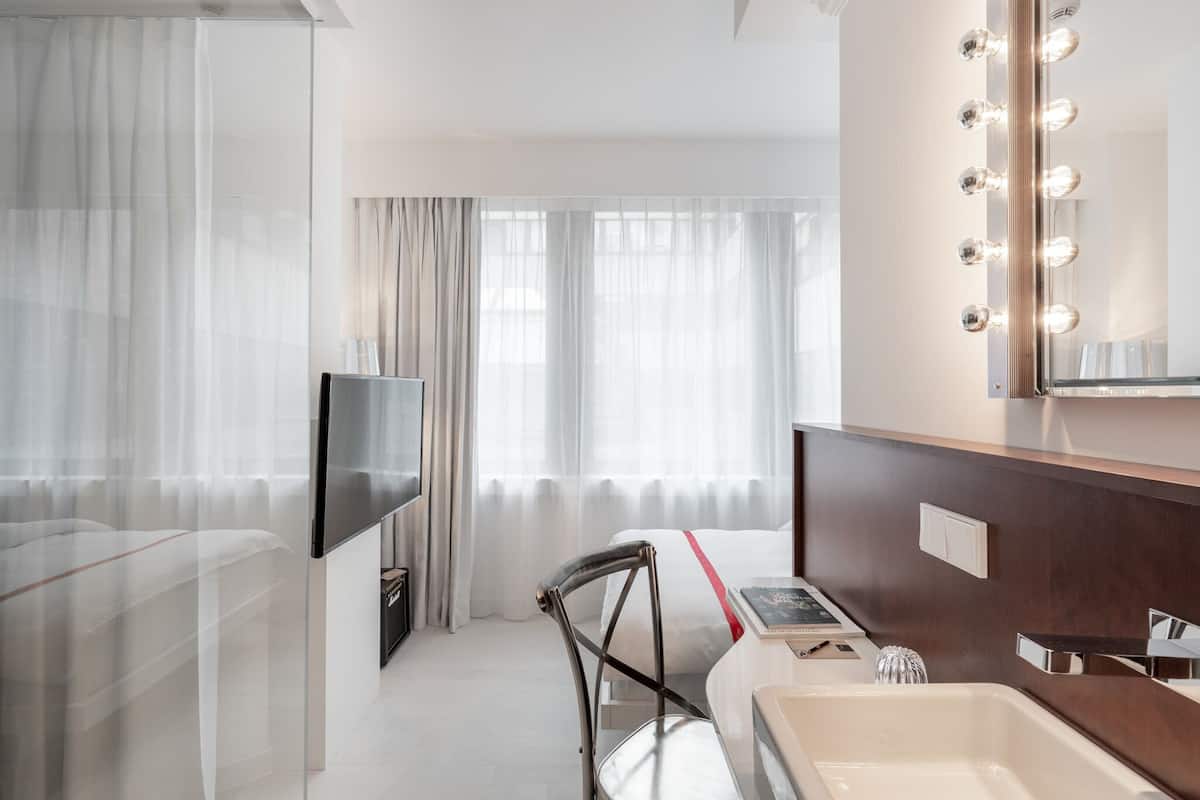 Una habitación de hotel moderna con una televisión de pantalla plana, una tocador con espejo e iluminación, una silla, una cama y un ventanal con cortinas traslúcidas.