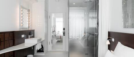 A modern hotel room with a large bed, a vanity with a mirror and lights, a bathroom with a shower, and a view into the adjacent room.
