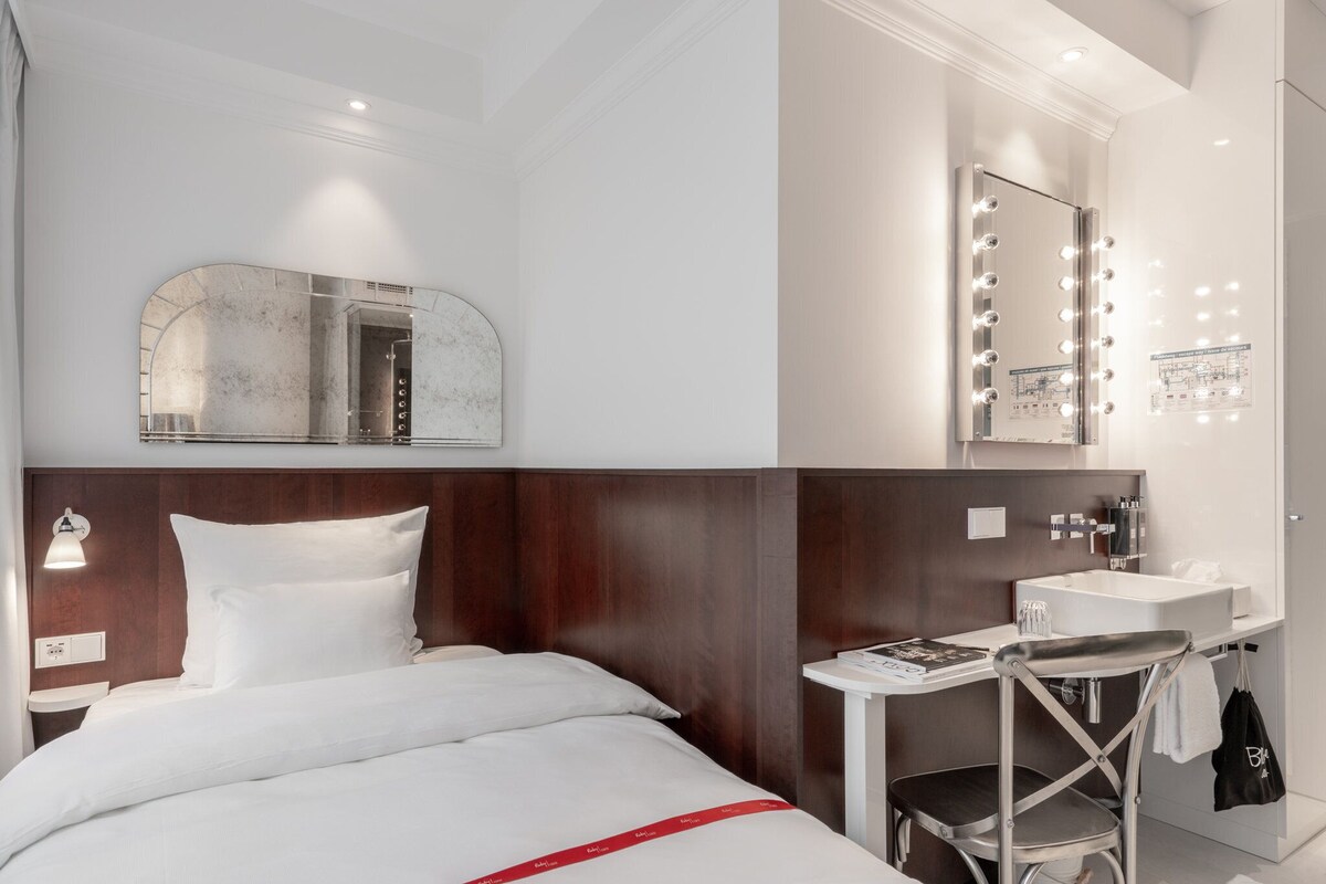 A hotel room with a bed, a vanity with a mirror and lights, a small table, and a chair.