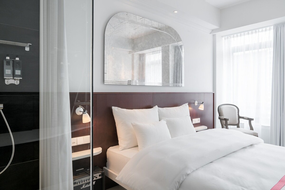 A hotel room with a bed, a large mirror, a chair, and a shower area.