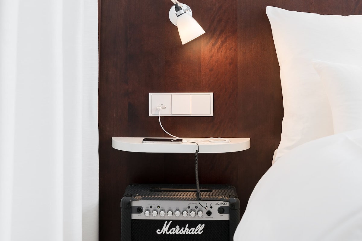 Una cama con ropa de cama blanca, un amplificador Marshall, una mesita de noche blanca con una lámpara y un enchufe de pared.
