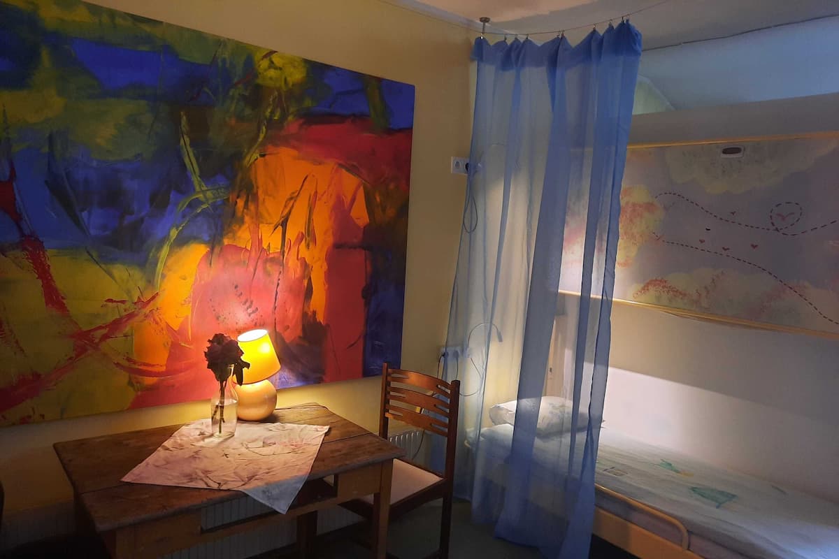 A room with a large abstract painting, a wooden table with a lamp, a chair, and a bed with a blue curtain.
