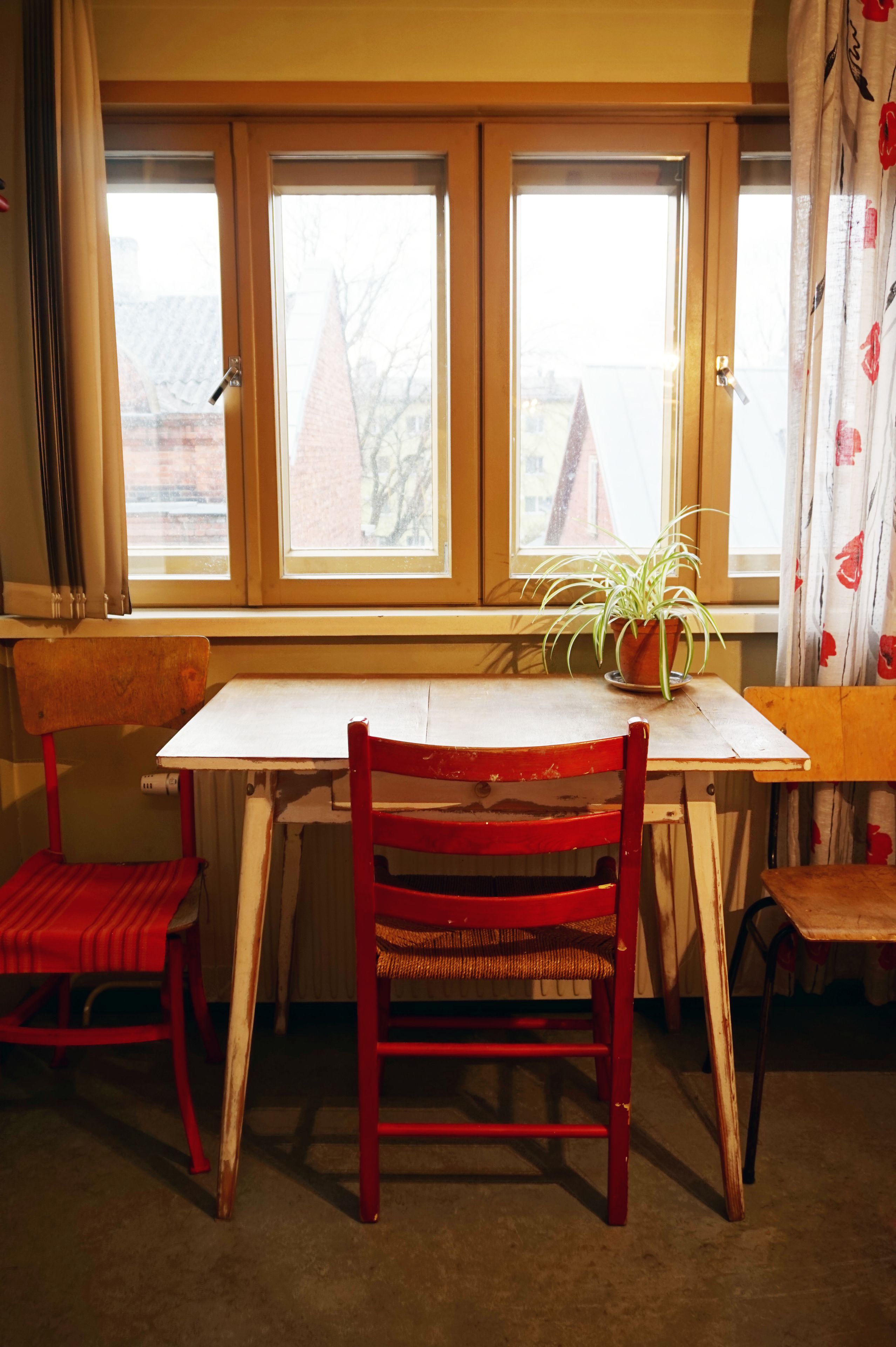 Una habitación con una mesa de madera y dos sillas rojas, una planta en maceta sobre la mesa, y una ventana con cortinas.