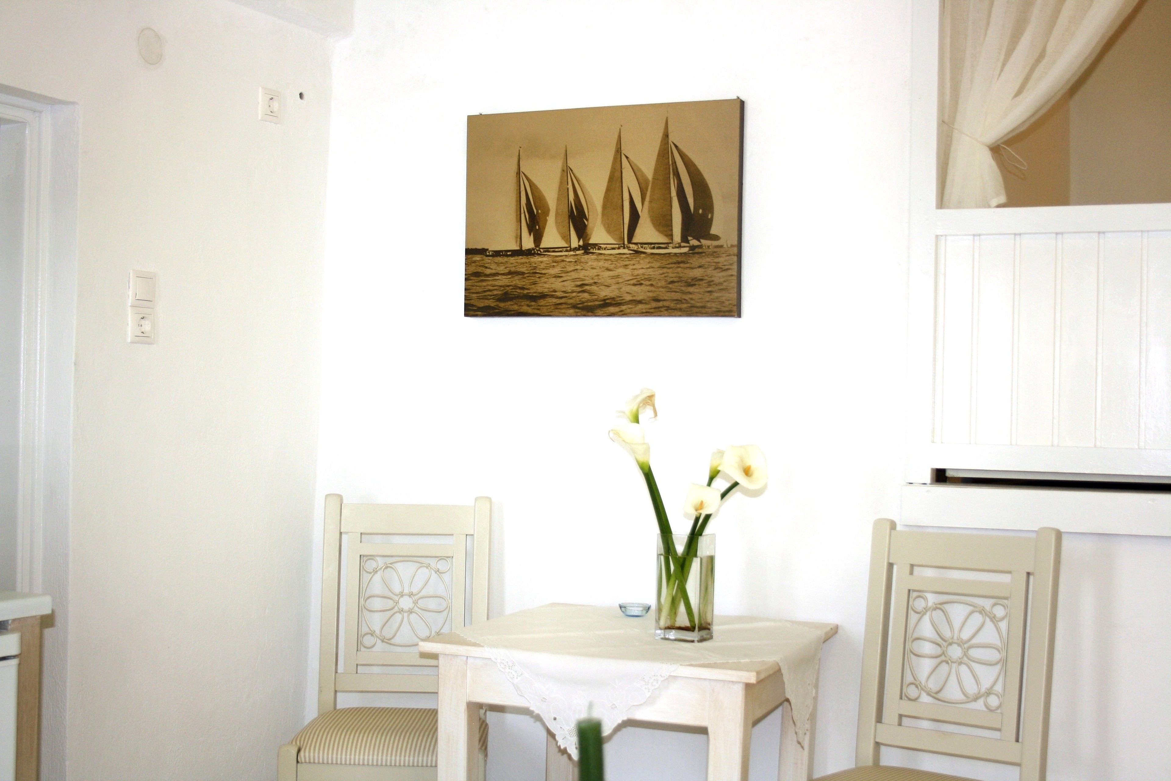 Een kamer met een witte muur, een schilderij van zeilboten in een lijst, een kleine tafel met een bloevas, en twee stoelen met decoratieve rugleuningen.