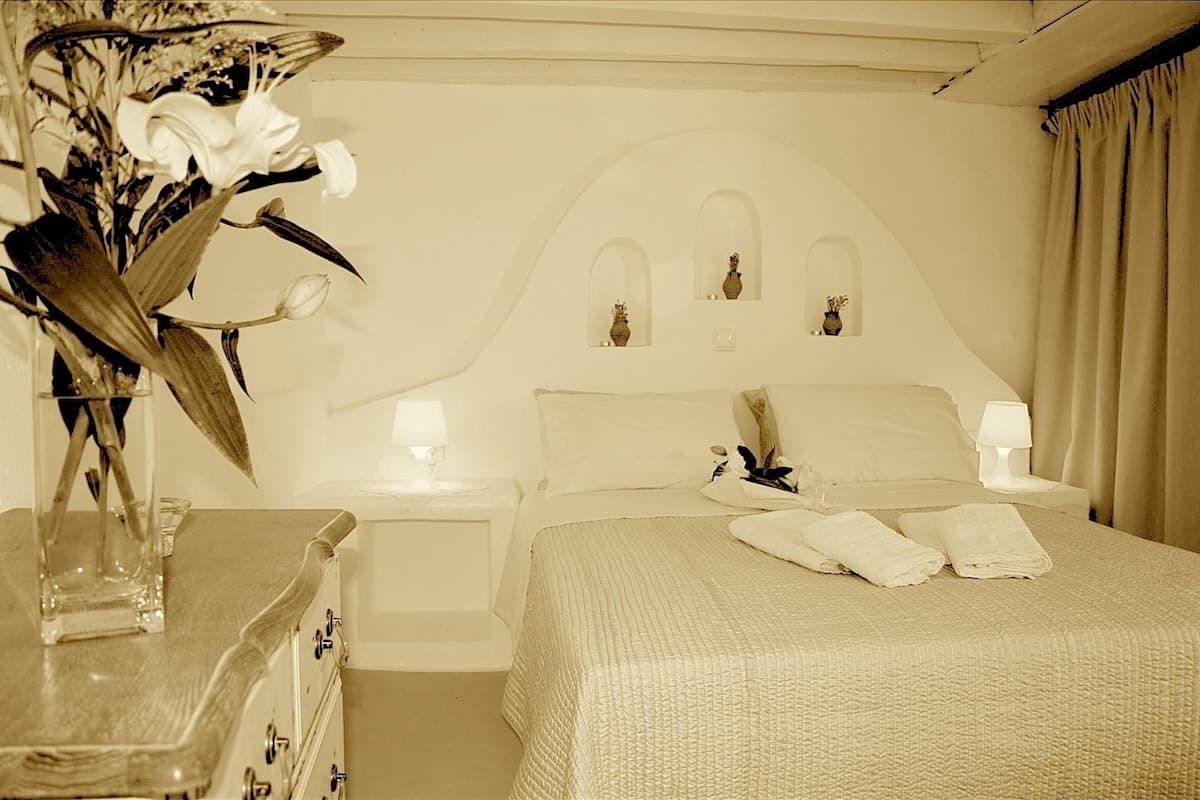 Une chambre d’hôtel comprenant un lit, deux tables de chevet, une petite table, un vase avec des fleurs et une commode.