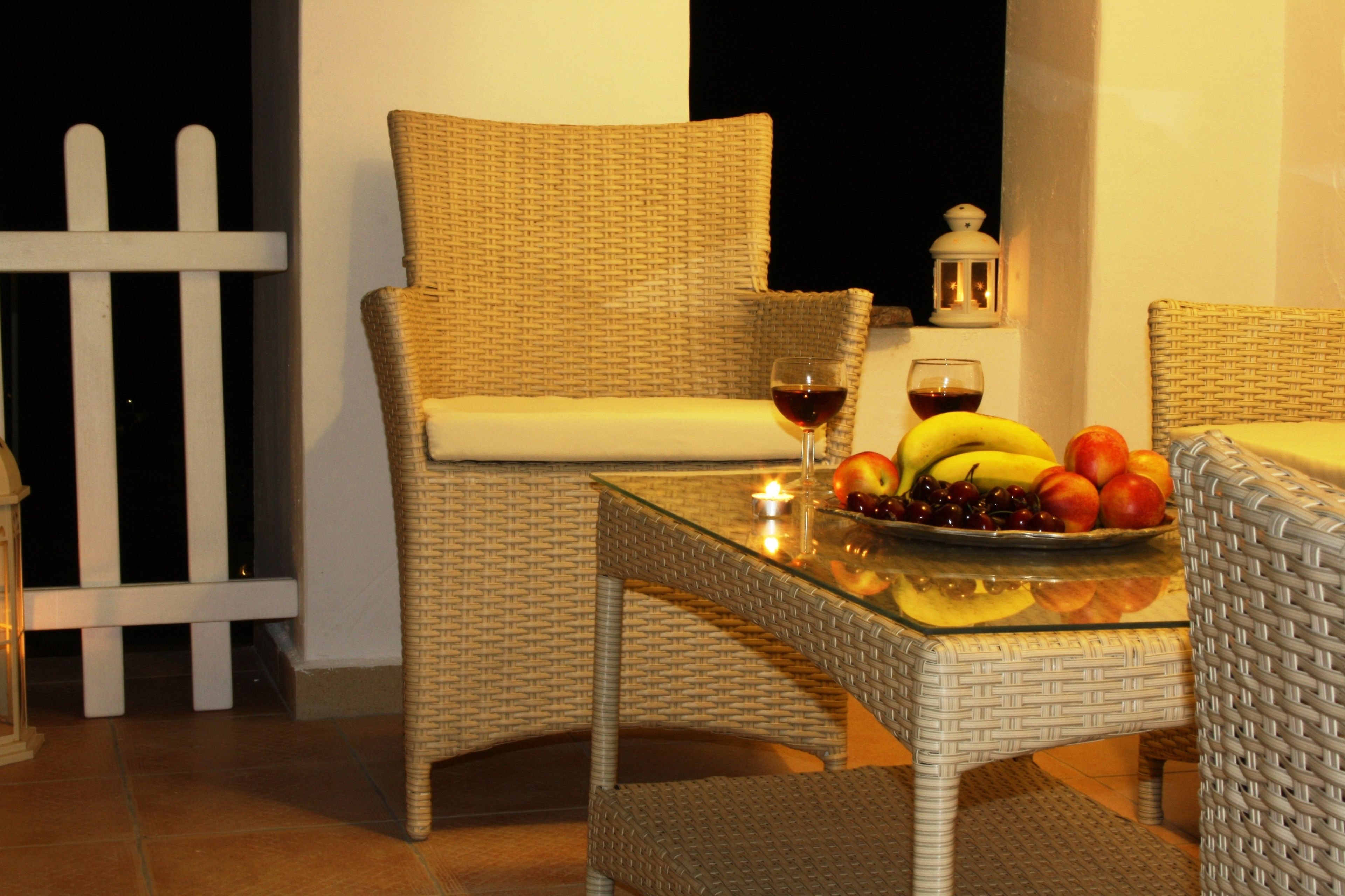 Una silla de mimbre con cojín, una mesa de vidrio con frutero y vela, y un farol blanco en un balcón.