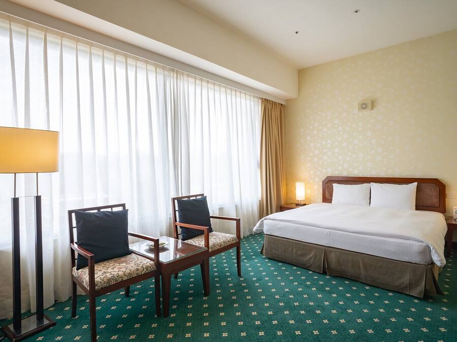 A hotel room with a bed, two chairs, a desk, a lamp, and a window with curtains.