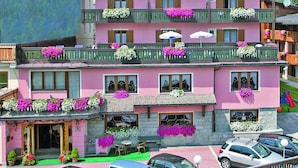 A multi-story hotel with balconies adorned with flowers, a stone facade, and a mountainous backdrop. - Alpen Hotel Vidi (Pinzolo)