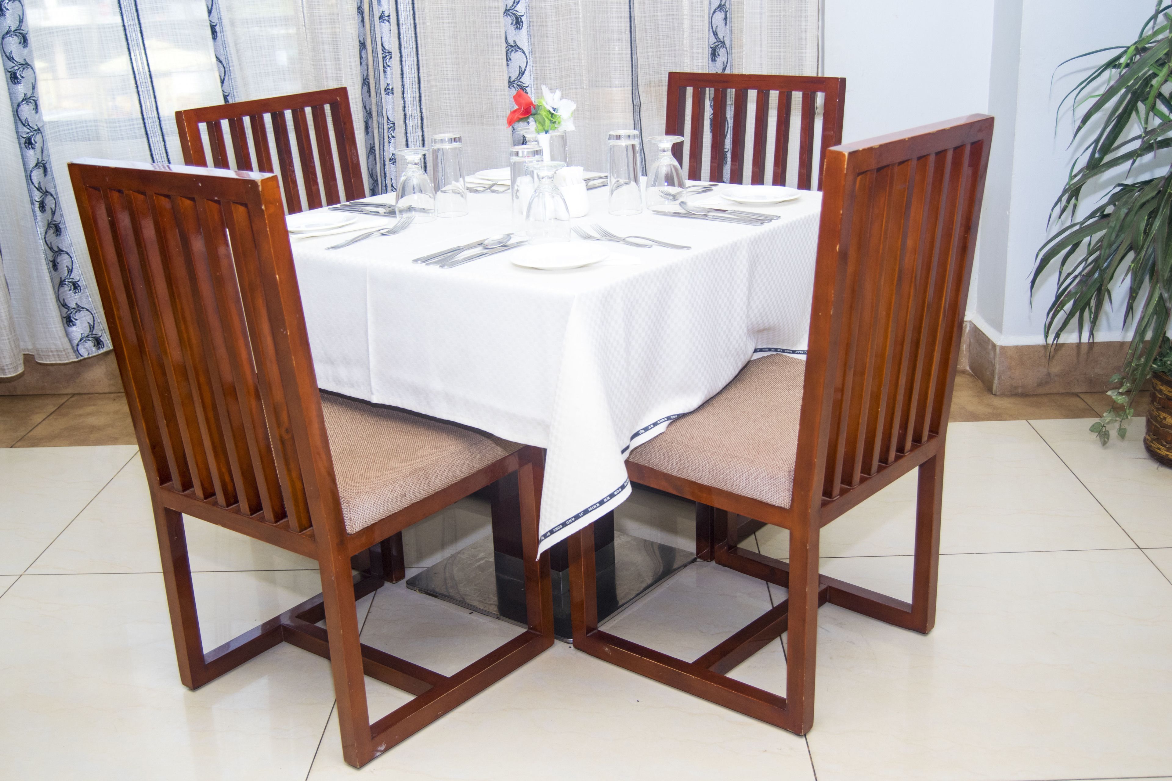 Una mesa de comedor preparada para cuatro personas, con sillas de madera, mantel blanco y un jarrón con flores.