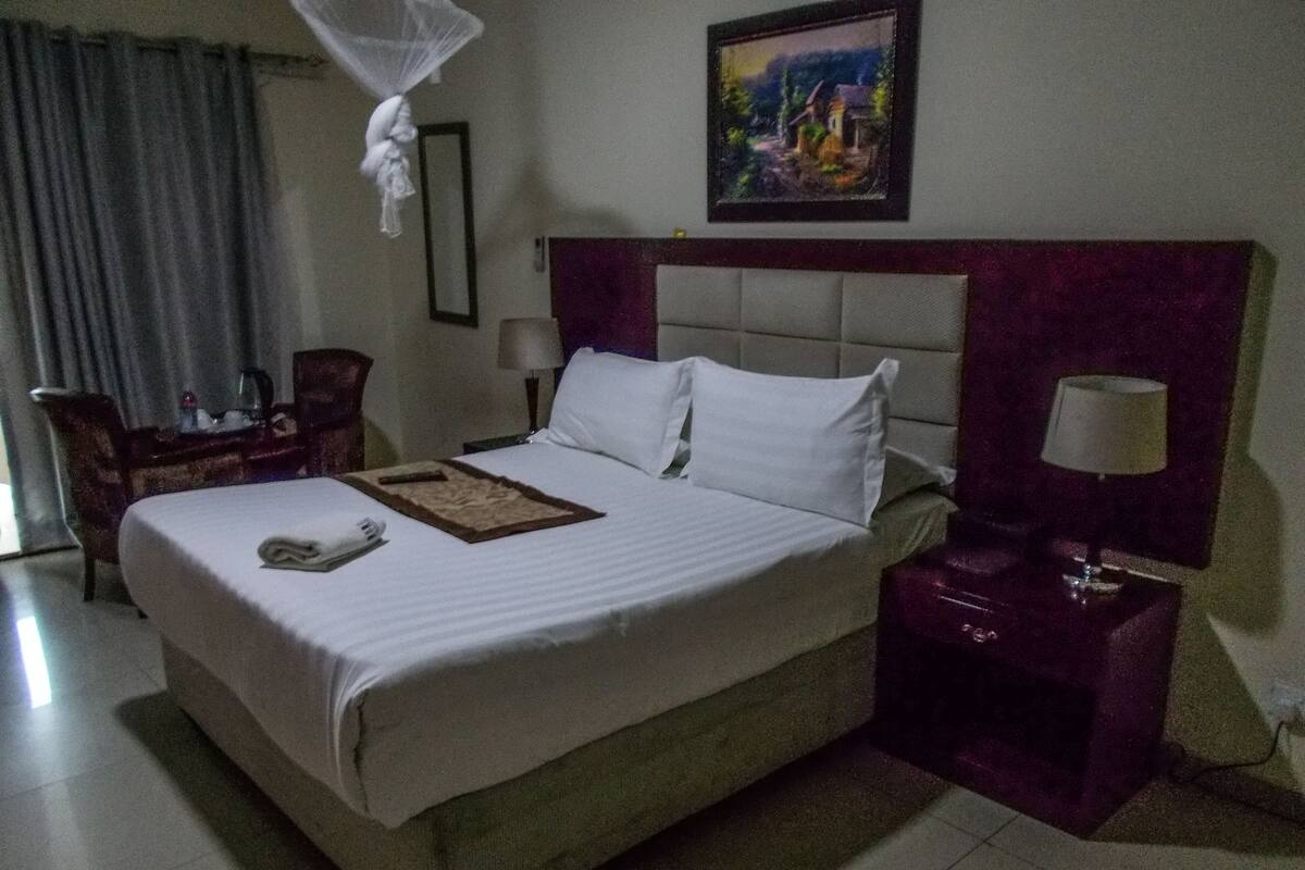 Quarto de hotel com uma cama grande, uma mesa de cabeceira com abajur, uma cadeira, uma escrivaninha com abajur e um quadro na parede.