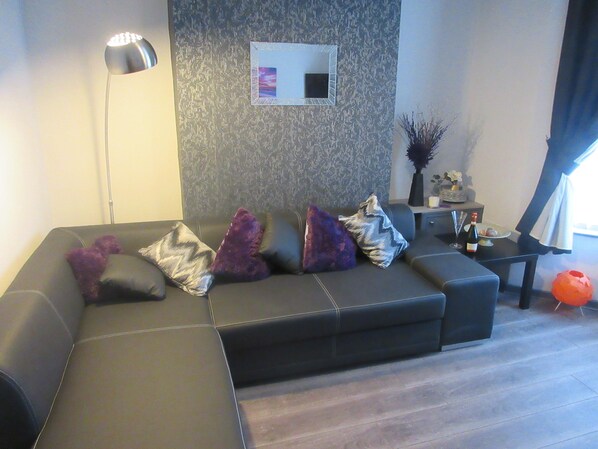 A modern living room with a sectional sofa, a floor lamp, a small table with a vase, and a decorative mirror on the wall.