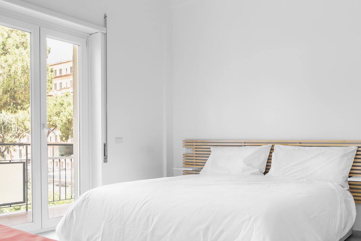 Un dormitorio con cama, cabecera de madera y vistas a un balcón con vegetación y edificios.