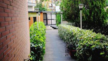 A narrow pathway flanked by brick walls and lush greenery, leading to a gated entrance with a small building in the background.