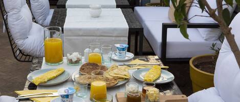 Una mesa de desayuno dispuesta con platos, vasos y cubiertos, rodeada de sillas blancas con cojines.