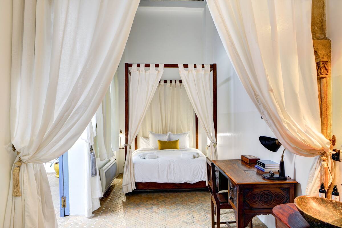 A four-poster bed with white linens and a yellow pillow, a wooden desk with a lamp, and a large woven basket.