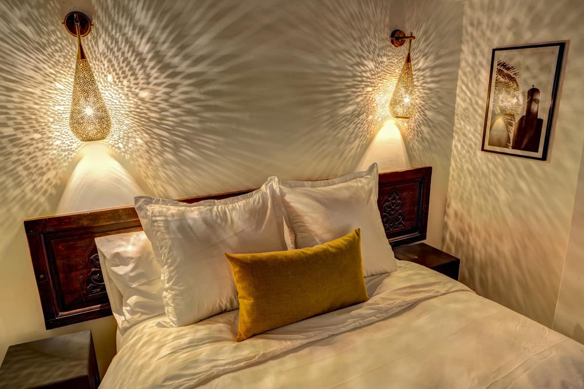 A neatly made bed with white and yellow pillows, two wall-mounted lamps, and a framed picture on the wall.