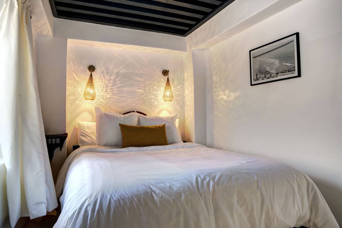 A bedroom with a bed, two wall-mounted lamps, a framed picture, and a curtain.