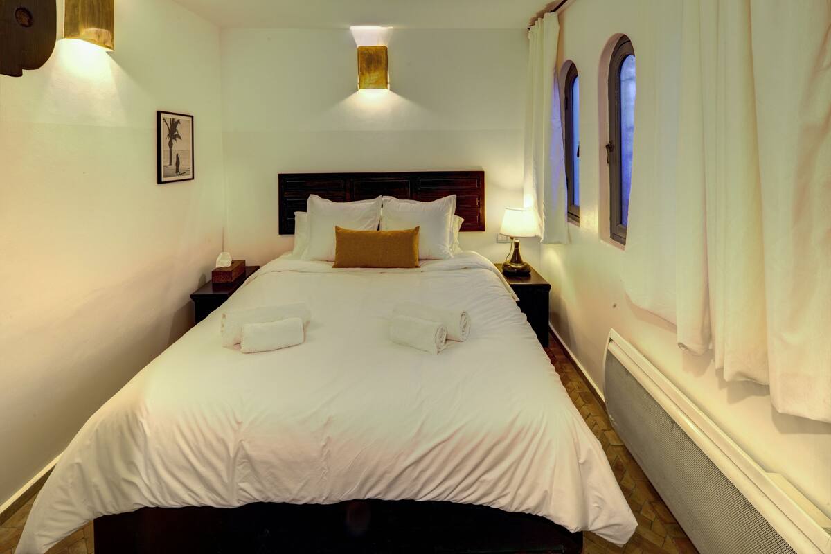 A neatly made bed with white linens and two towels, a nightstand with a lamp, and a framed picture on the wall.