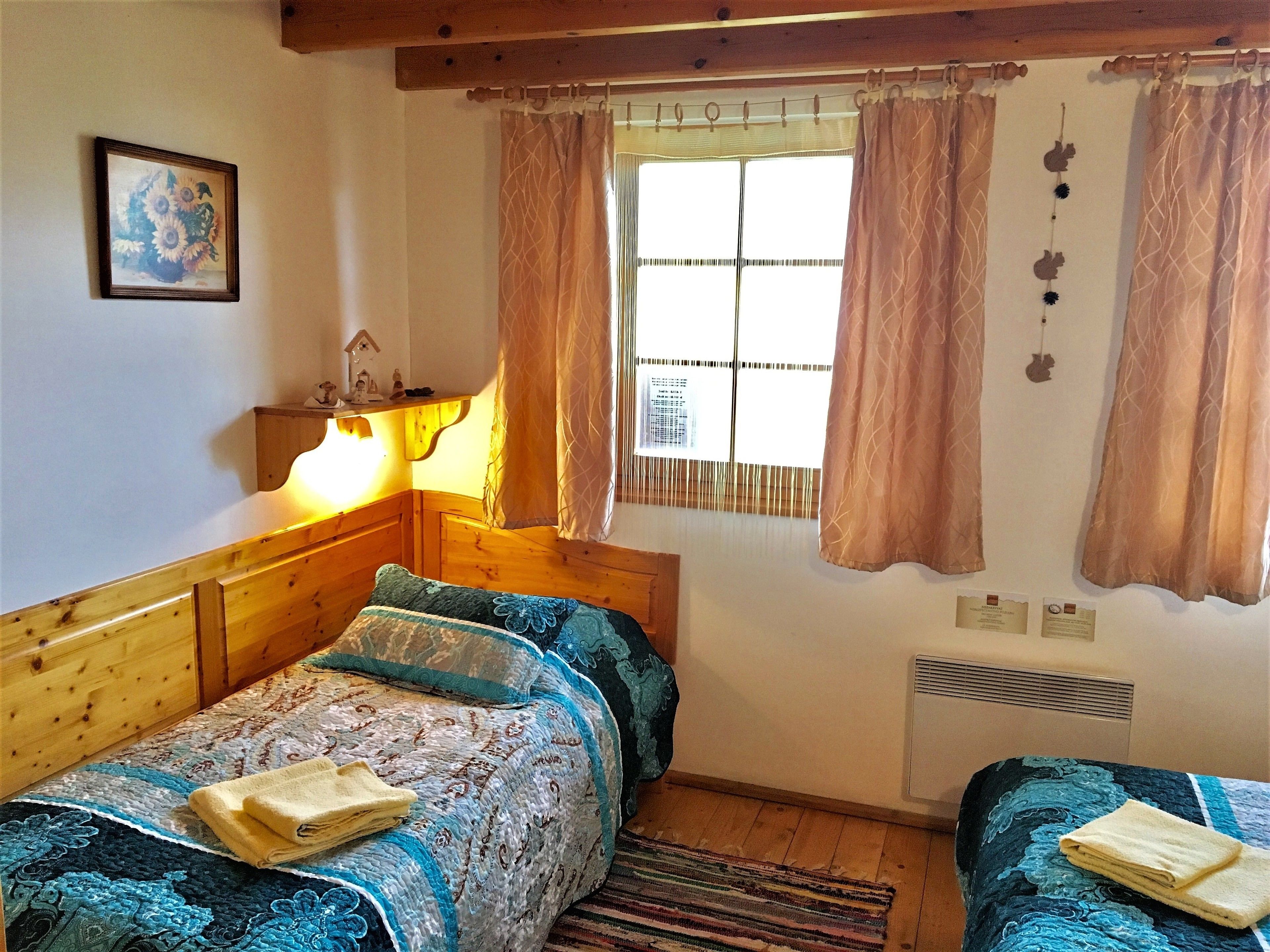 A cozy room with two beds, a wooden headboard, a window with curtains, a radiator, and a small shelf with decorative items.