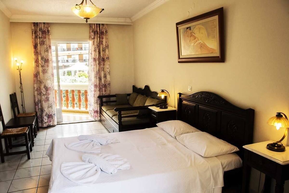 A hotel room with a bed, a desk, a chair, a nightstand, a lamp, a painting, and a view of the outside balcony.