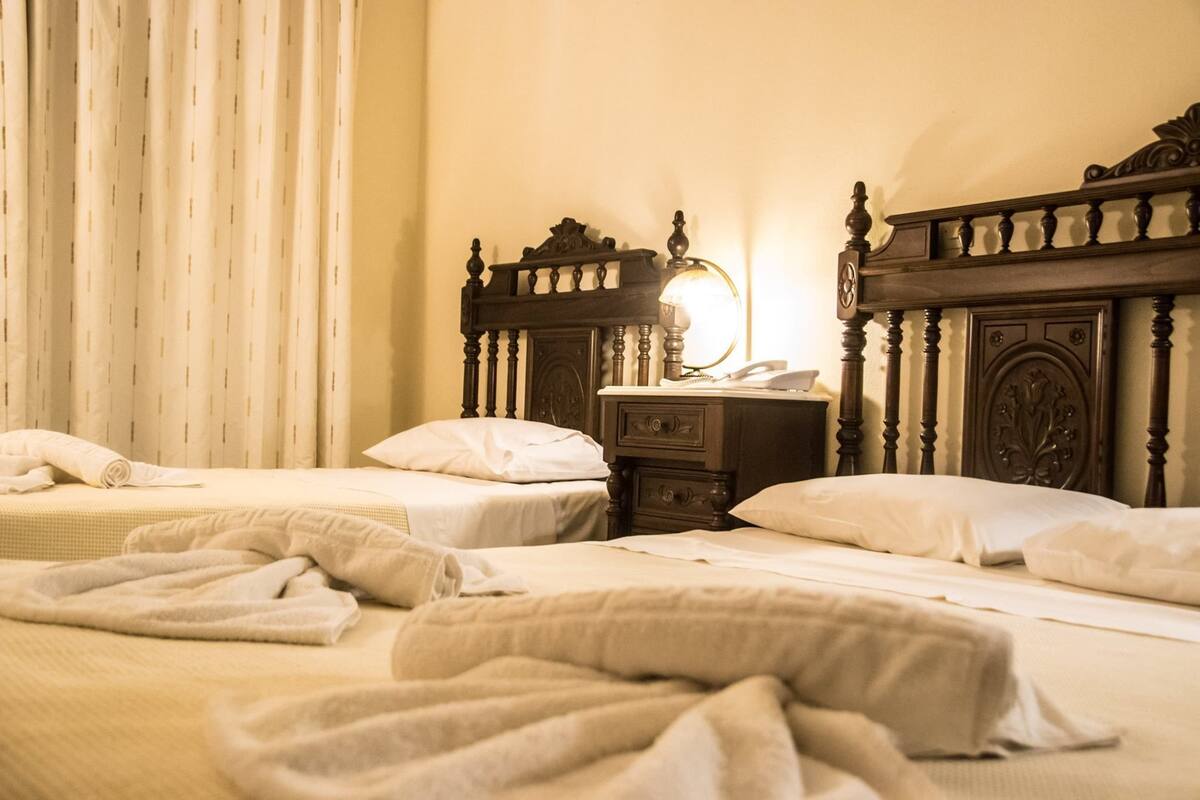 A hotel room with two beds, a nightstand, a lamp, and a window with curtains.