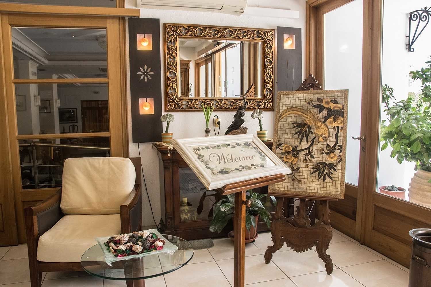 A reception area with a leather chair, a glass table with snacks, a welcome sign, a mirror with an ornate frame, and a decorative wall piece.