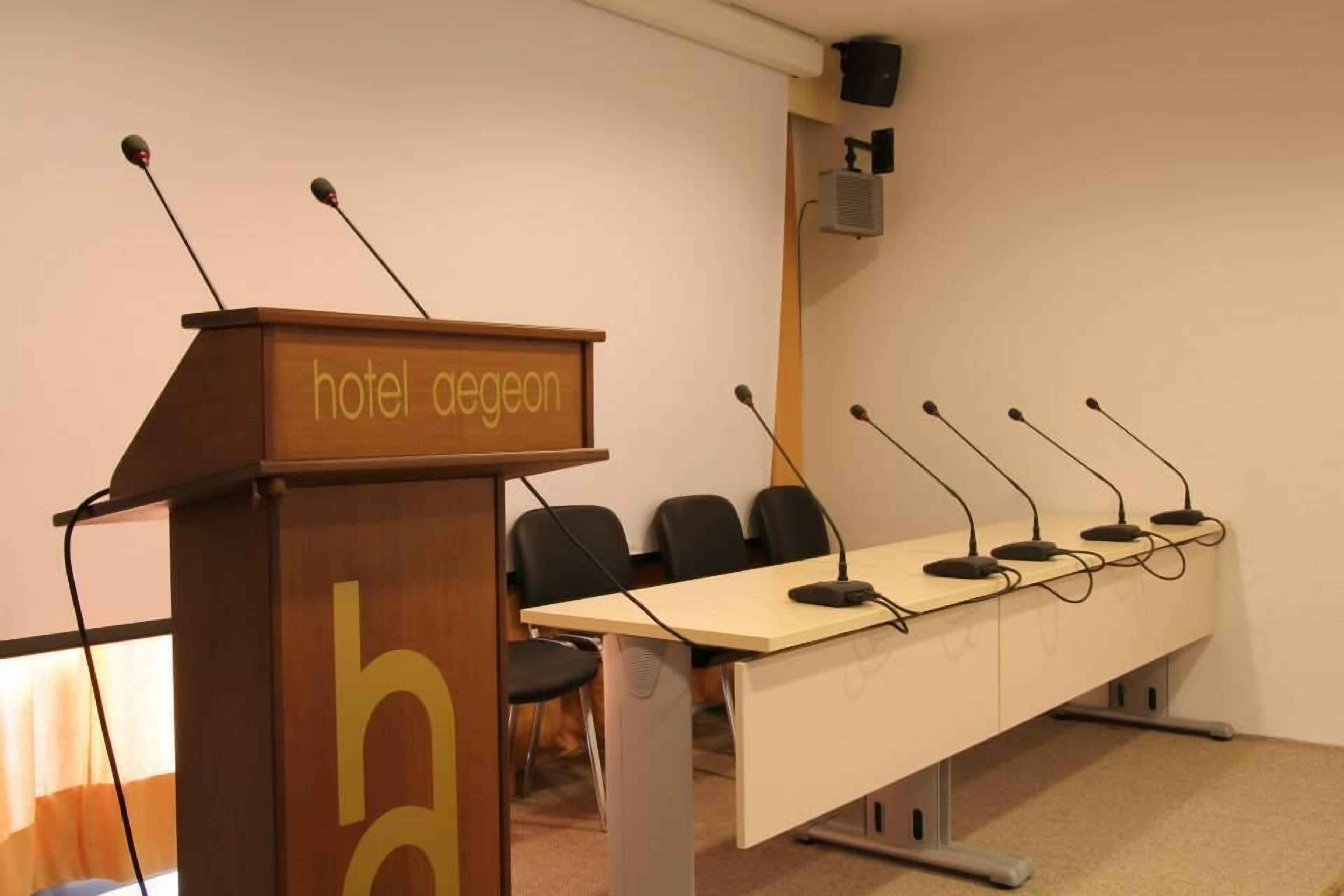 A conference room with a podium labeled "hotel aegeon" and a long table with multiple microphones.
