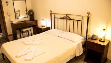 A neatly made bed with white linens, a bedside table with a lamp, a small desk with a mirror, and a trash can.