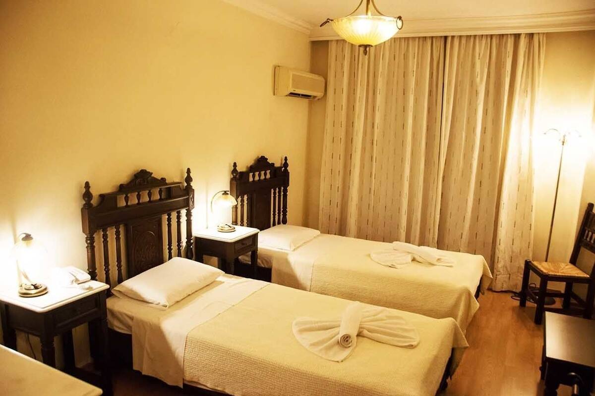 A hotel room with two single beds, a bedside table with a lamp, a chair, and a large window with curtains.