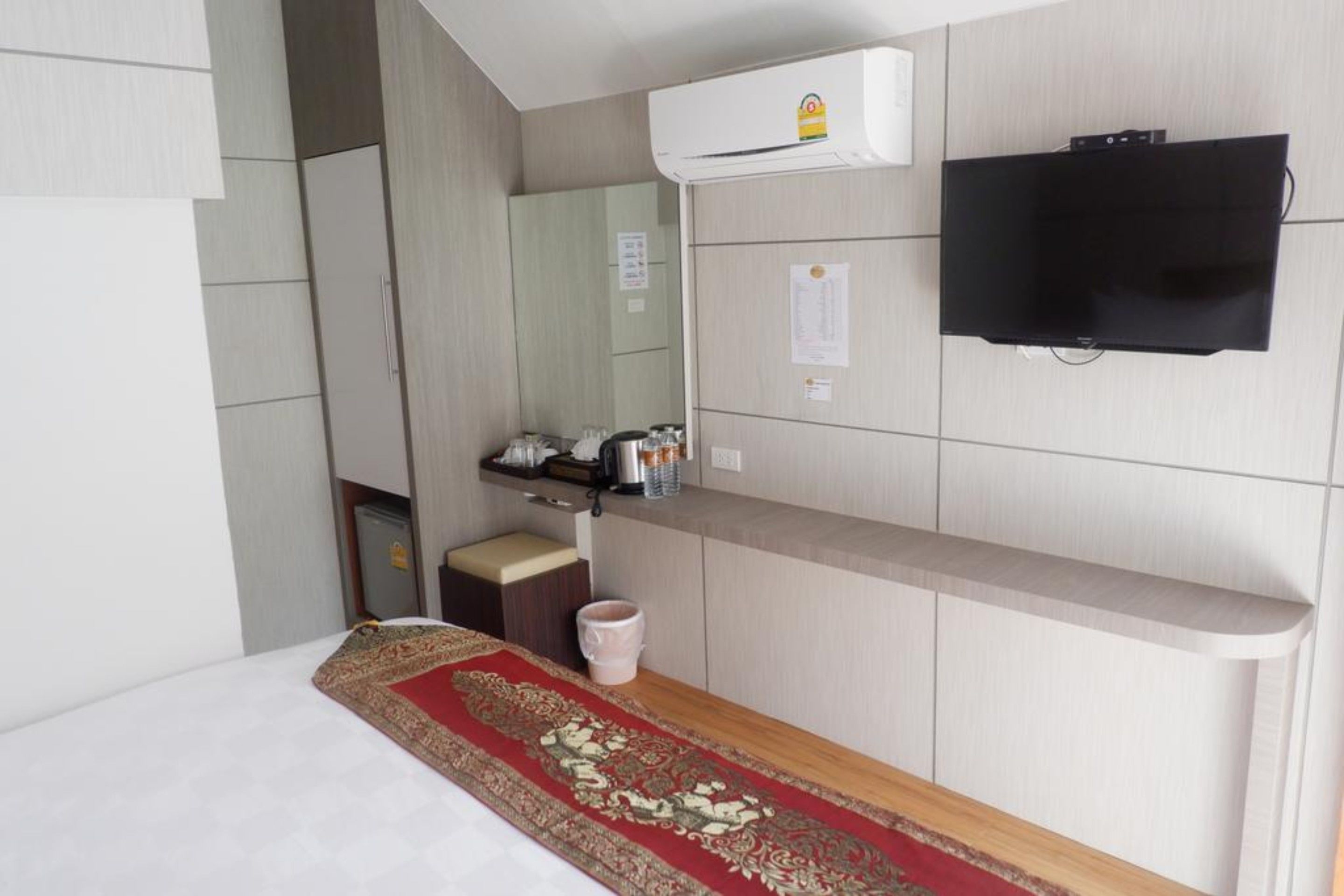 A hotel room with a bed, a red patterned runner, a television mounted on the wall, a small table with a kettle, and a cabinet with a mirror.