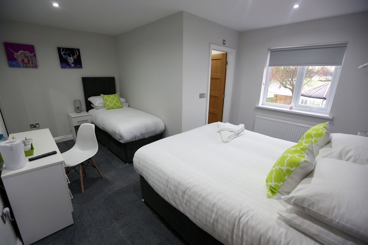 A hotel room with two single beds, a desk with a chair, a small cabinet, and a window with a view of the outside.