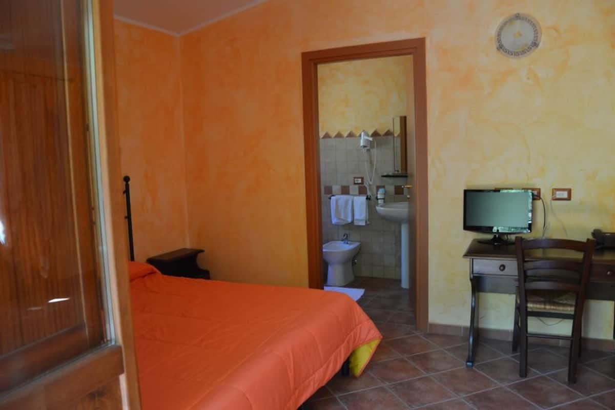 Un dormitorio con cama naranja, un escritorio con computadora, una silla, una ventana y un baño visible a través de una puerta abierta.