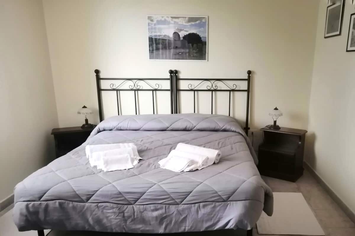 Una cama con colcha gris, dos toallas blancas, un cabecero de metal y dos mesitas de noche con lámparas.