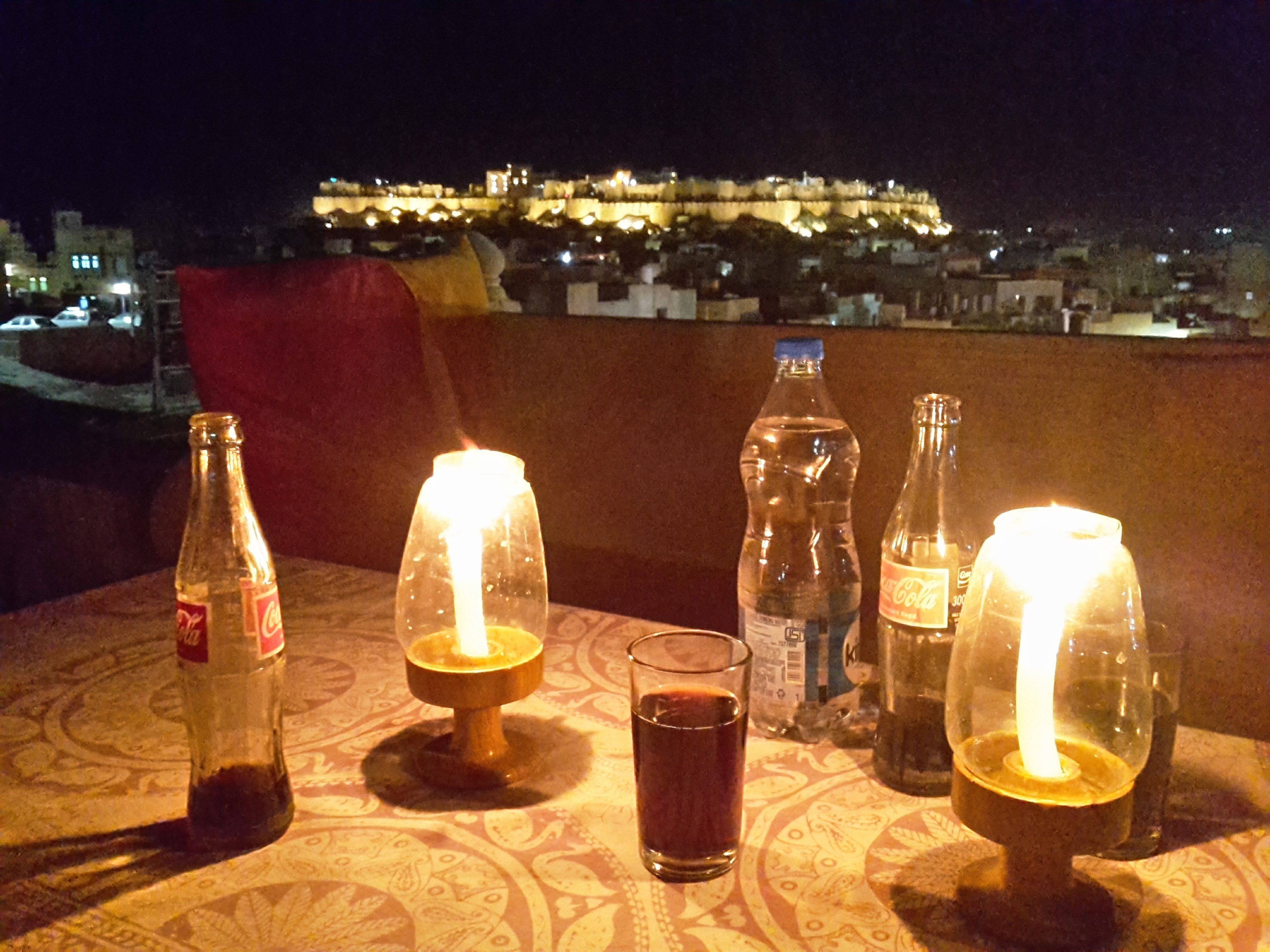 A table with bottles, glasses, and a candle, with a cityscape and castle in the background.