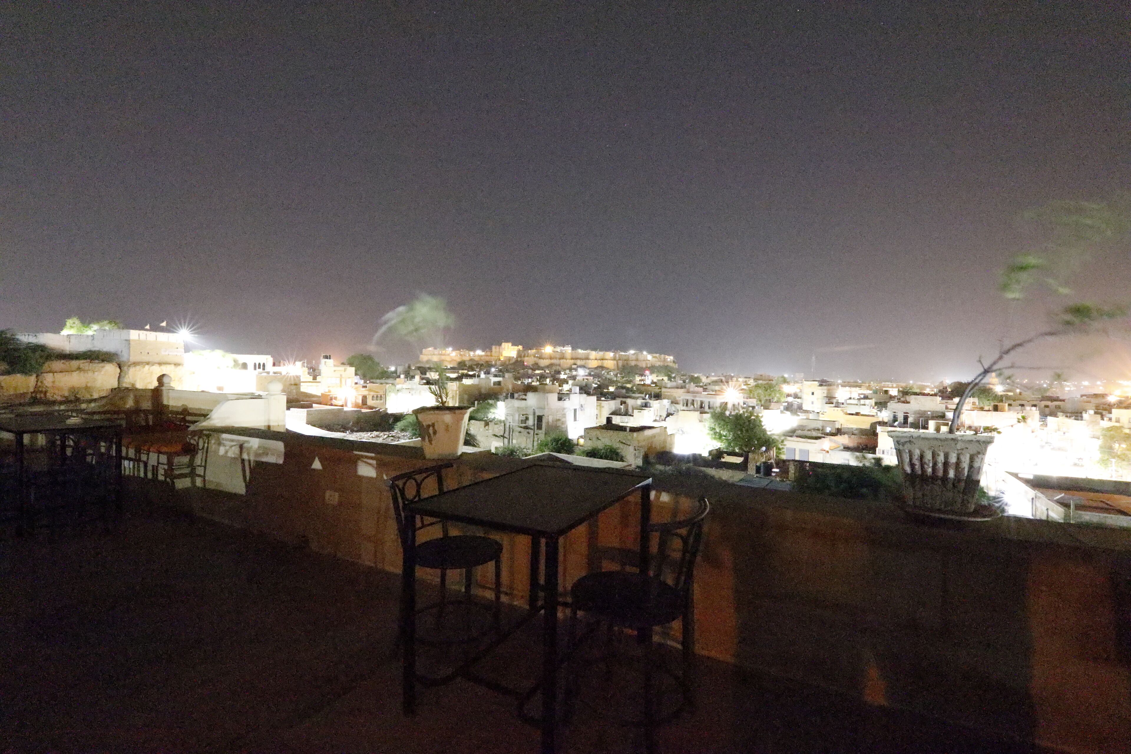 A rooftop terrace with a table and chairs, overlooking a cityscape at night.