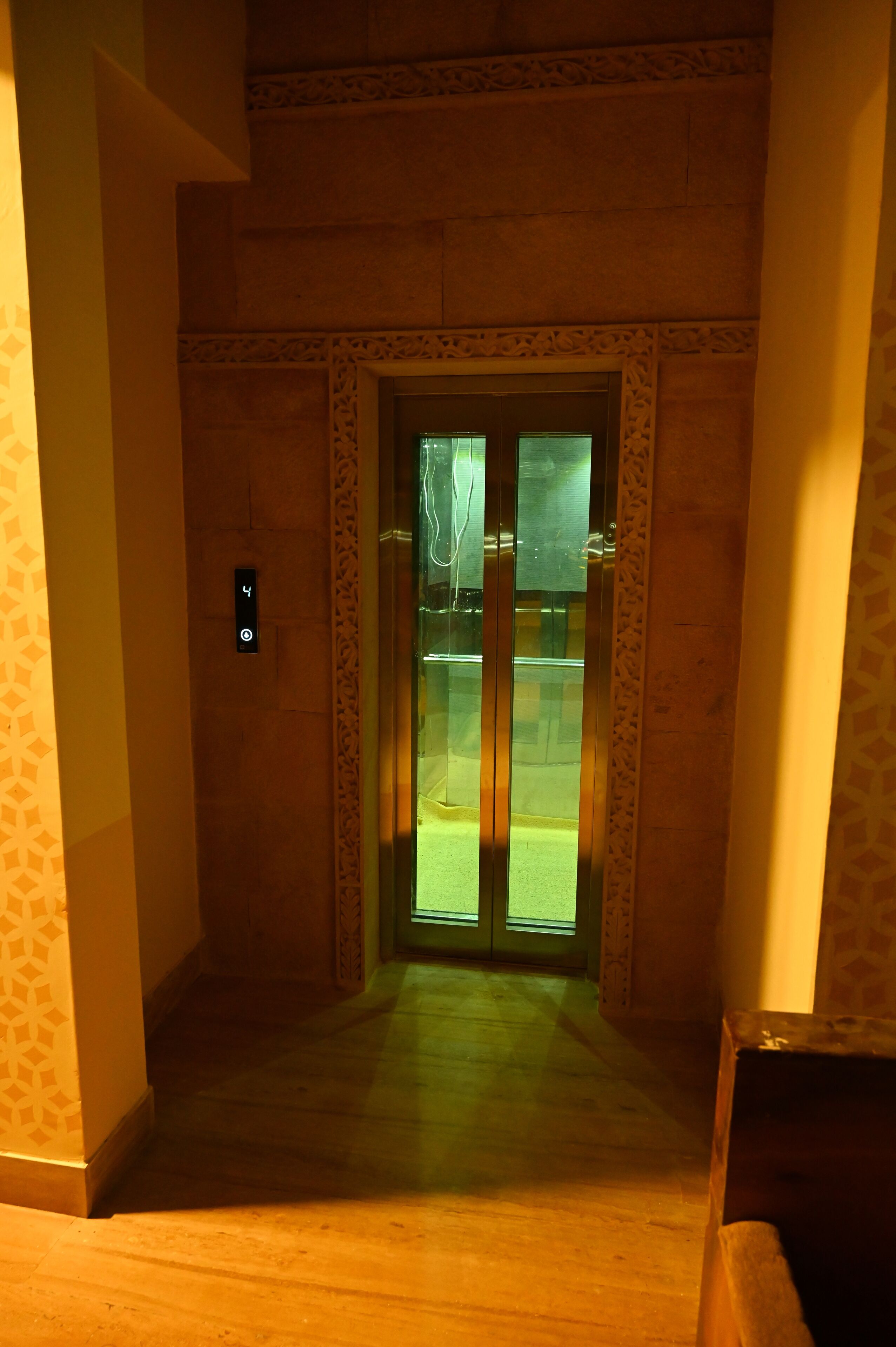 A narrow hallway with a wooden floor and a glass elevator door with a digital display.