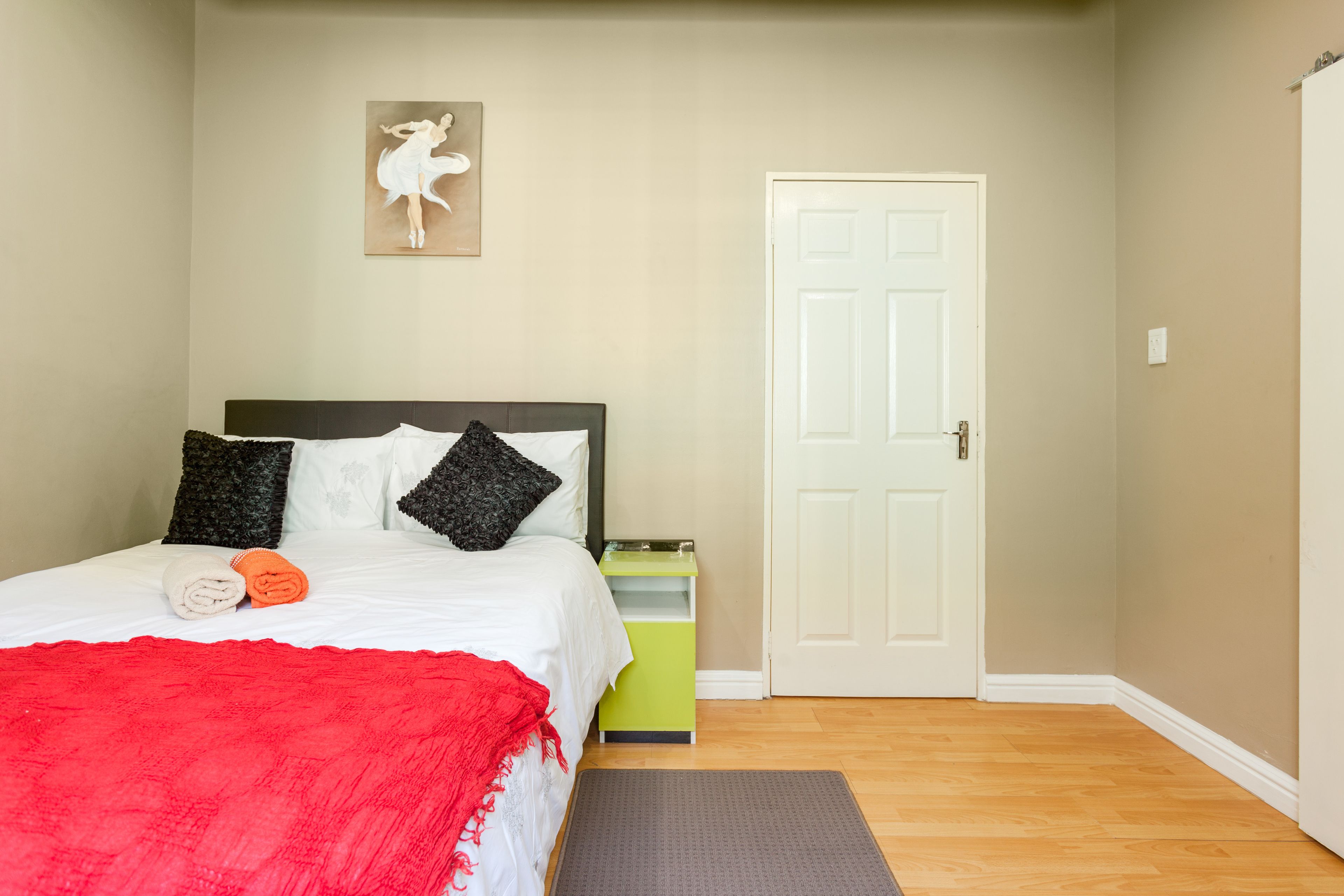 Une chambre à coucher avec un lit, des oreillers, une couverture rouge, une table de chevet, une porte et un tableau accroché au mur.