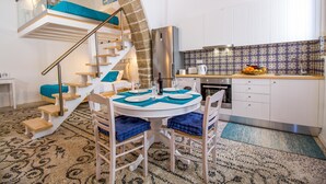 A compact kitchen with white cabinets, a stainless steel refrigerator, and a dining area with a round table and blue cushions. - The Red Door in Koskinou (Rhodes)