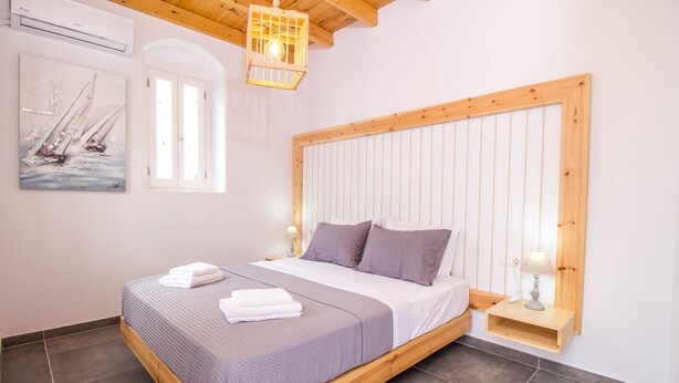 A bedroom with a wooden bed frame, a headboard with vertical slats, a bedside table with a lamp, and a painting of sailboats on the wall. - The Red Door in Koskinou (Rhodes)