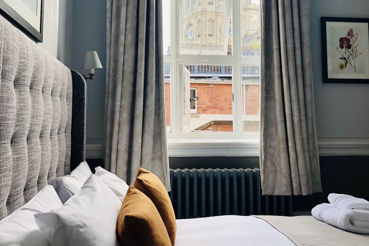 Quarto de hotel com uma janela grande, uma cama com almofadas brancas e castanhas, um radiador e um quadro emoldurado na parede.