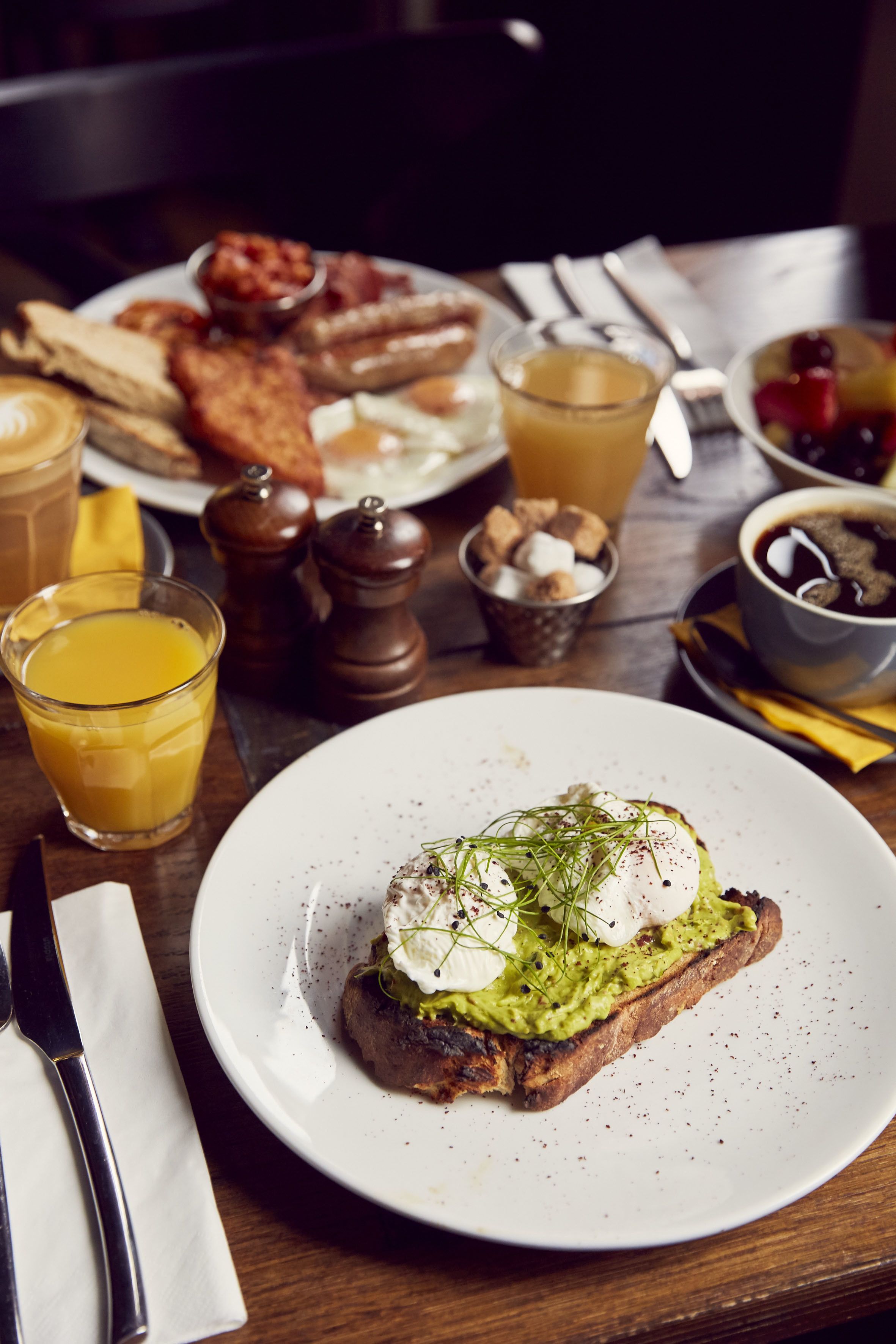 Un desayuno con tostadas cubiertas de aguacate y huevos pochados, un plato de tocino y huevos, un vaso de jugo de naranja y una taza de café.