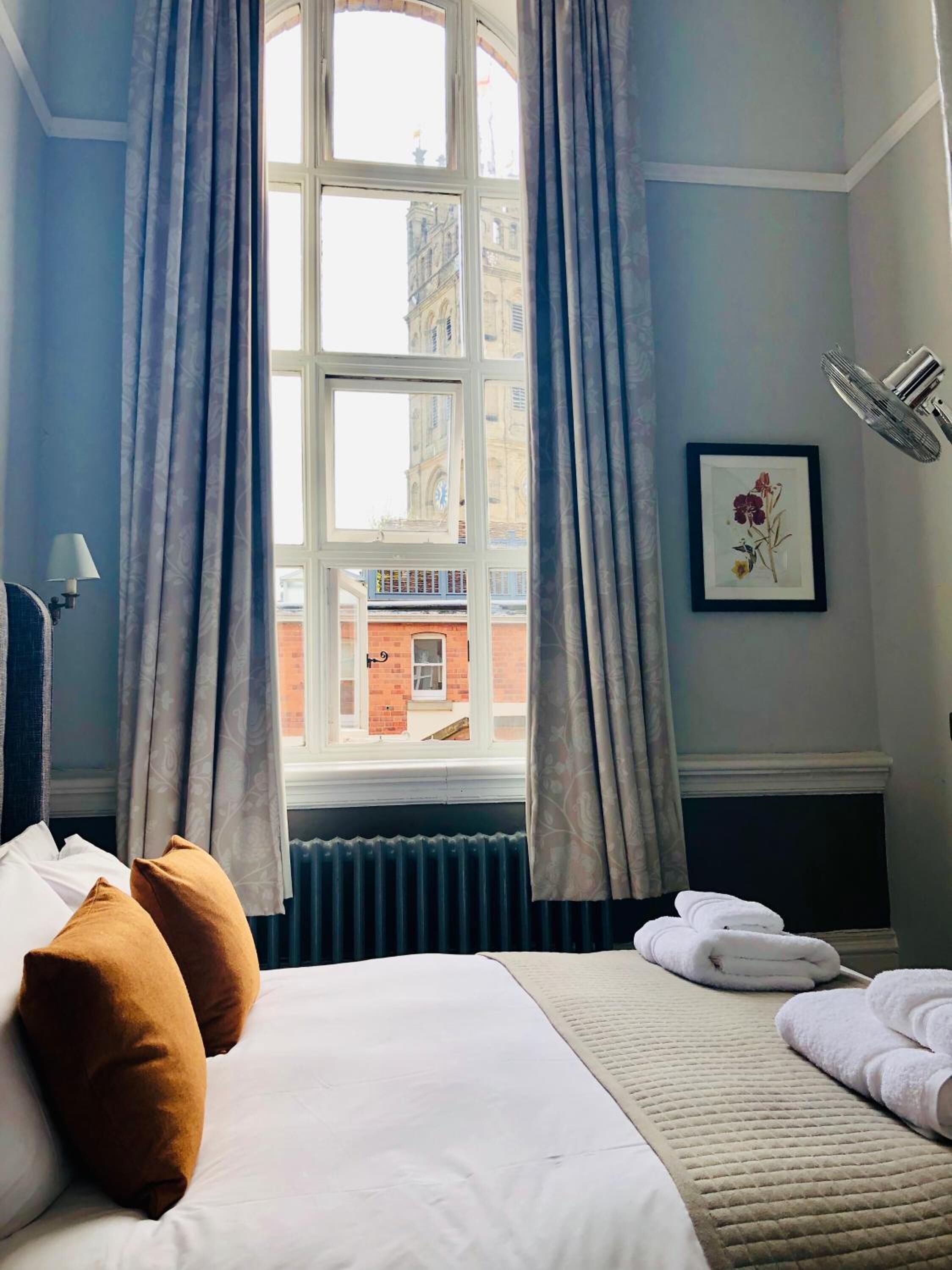 A hotel room with a large window, a bed with white linens, brown pillows, a bedside table with a lamp, a framed picture on the wall, and a radiator below the window.