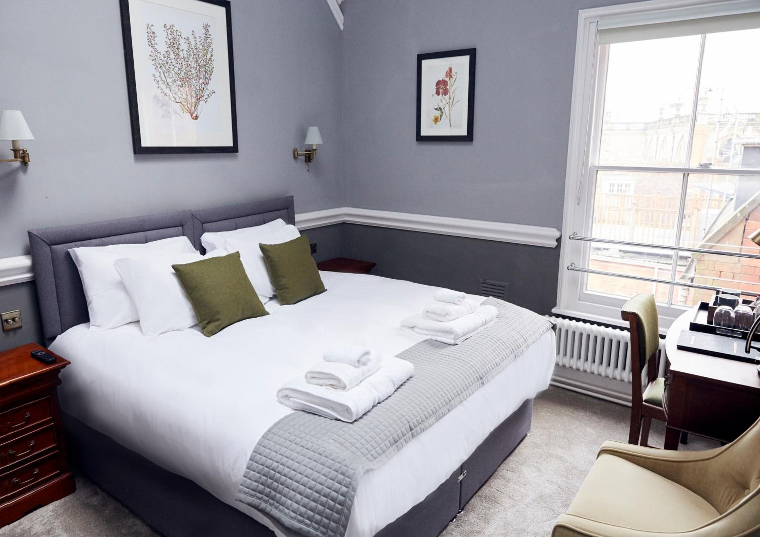 A neatly made bed with white linens and a grey quilt, a wooden nightstand with a remote control, and a chair in a room with grey walls and framed botanical prints.